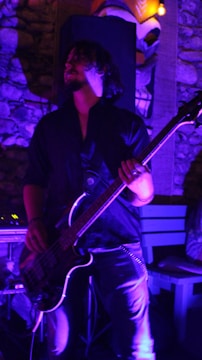 A person with long hair is playing an electric bass guitar under dim, purple lighting. They are wearing a dark outfit, standing in front of a stone wall with some wooden elements, giving the scene an intimate and relaxed atmosphere. The focus is on the lower half of their face and the guitar.