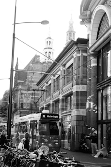 A busy urban street scene featuring an electric tram labeled '16 Wateringen' moving along tracks. The surrounding architecture includes historic buildings with ornate details and tall church spires in the background. Numerous bicycles are parked in the foreground, and several pedestrians are walking or standing nearby.
