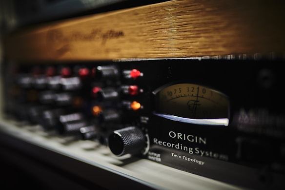 A close-up image of a professional audio recording system with various control knobs, switches, and a display panel. The panel includes a VU meter with numbers and a needle indicator. The equipment piece is labeled with 'ORIGIN Recording System' and 'Twin Topology.' The materials appear to include metal and wood.