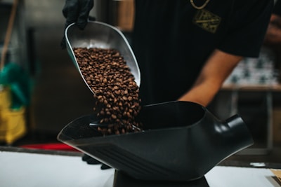 A person wearing a black glove is pouring roasted coffee beans from a metal scoop into a black container. The background is blurred, suggesting a setting related to coffee preparation or production.