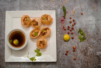 Five crispy puris filled with savory ingredients are placed on a square white plate. A small bowl of tangy water with a lemon slice floats nearby. Scattered around are a green chili, peanuts, a yellow lemon, and sprigs of coriander, creating a rustic presentation.