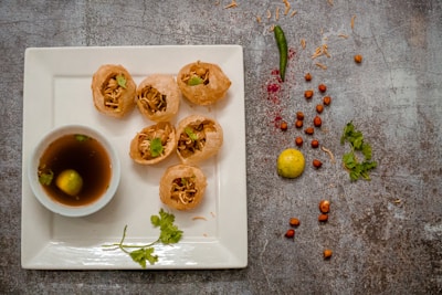 Five crispy puris filled with savory ingredients are placed on a square white plate. A small bowl of tangy water with a lemon slice floats nearby. Scattered around are a green chili, peanuts, a yellow lemon, and sprigs of coriander, creating a rustic presentation.