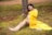 A pregnant woman sits on the ground, leaning against a tree, wearing a flowing yellow dress. She is sitting in a grassy area with scattered red petals and appears to be gently cradling her belly. Beside her are a pair of small white baby shoes placed on the grass.
