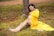 A pregnant woman sits on the ground, leaning against a tree, wearing a flowing yellow dress. She is sitting in a grassy area with scattered red petals and appears to be gently cradling her belly. Beside her are a pair of small white baby shoes placed on the grass.