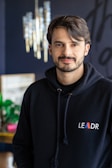 A person with short dark hair and a beard is wearing a black hoodie with the word 'LEADR' on it. They are indoors, with a modern chandelier visible in the background. The environment appears stylish and contemporary, with some greenery and blurred decorative elements.
