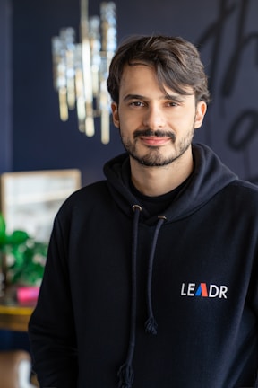 A person with short dark hair and a beard is wearing a black hoodie with the word 'LEADR' on it. They are indoors, with a modern chandelier visible in the background. The environment appears stylish and contemporary, with some greenery and blurred decorative elements.