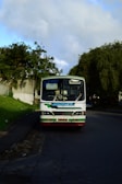 A white bus with a sign reading 'Contract Bus' is parked on the side of a quiet road. It is surrounded by greenery, with large trees and bushes nearby. The sky is partly cloudy, suggesting a calm day.