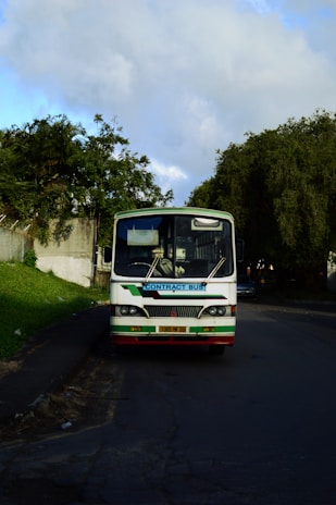A white bus with a sign reading 'Contract Bus' is parked on the side of a quiet road. It is surrounded by greenery, with large trees and bushes nearby. The sky is partly cloudy, suggesting a calm day.