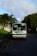 A white bus with a sign reading 'Contract Bus' is parked on the side of a quiet road. It is surrounded by greenery, with large trees and bushes nearby. The sky is partly cloudy, suggesting a calm day.