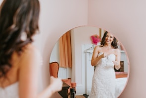A woman in a wedding dress appears in a mirror, smiling and possibly adjusting her attire. The room has a warm, soft ambiance with peach-colored walls and some pink decorative elements visible in the background.