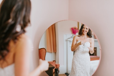 A woman in a wedding dress appears in a mirror, smiling and possibly adjusting her attire. The room has a warm, soft ambiance with peach-colored walls and some pink decorative elements visible in the background.