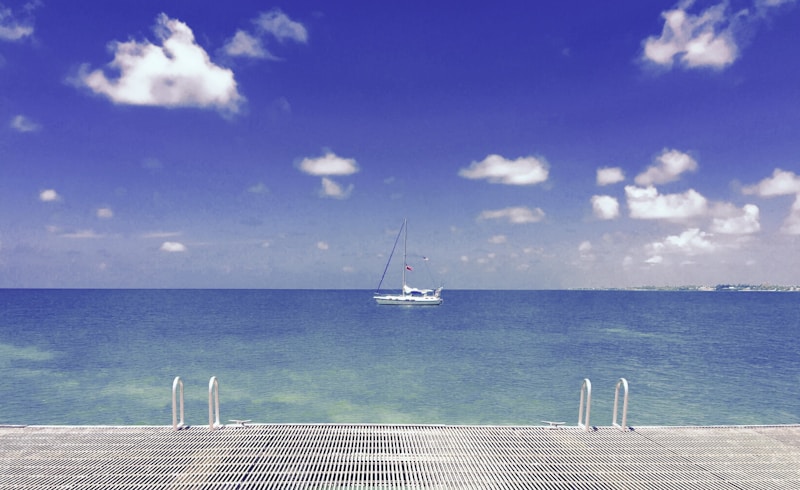 A serene seascape featuring a sailboat on calm, turquoise waters beneath a clear blue sky dotted with fluffy white clouds. In the foreground, a metal deck with ladders reaching into the sea suggests a place for people to step into the water.