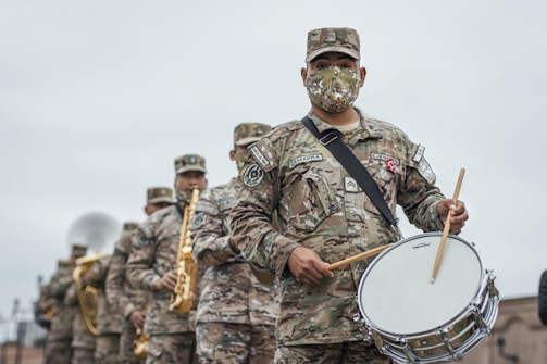 A group of military personnel in camouflage uniforms, with the foreground individual holding a snare drum and drumsticks. They are aligned in a procession, with visible musical instruments, including saxophones and a sousaphone. The sky is overcast, suggesting a somber or serious occasion.