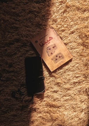 A book with Arabic text on the cover lies on a textured, golden carpet. Next to it, there is a black device, possibly a phone or notebook, with a cord wrapped around it. Sunlight casts soft shadows, creating a warm and cozy atmosphere.