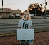 A person is standing on a brick sidewalk, holding a sign that reads 'JUSTICIA PARA VANESSA.' They are wearing a mask and a New York Yankees T-shirt. Behind them, there is a road with parked cars, and a store with a sign that says 'Locke Supply' is visible. The sky appears to be at sunset, with trees and streetlights in the background.