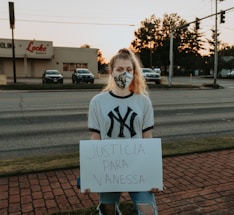 A person is standing on a brick sidewalk, holding a sign that reads 'JUSTICIA PARA VANESSA.' They are wearing a mask and a New York Yankees T-shirt. Behind them, there is a road with parked cars, and a store with a sign that says 'Locke Supply' is visible. The sky appears to be at sunset, with trees and streetlights in the background.