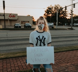 A person is standing on a brick sidewalk, holding a sign that reads 'JUSTICIA PARA VANESSA.' They are wearing a mask and a New York Yankees T-shirt. Behind them, there is a road with parked cars, and a store with a sign that says 'Locke Supply' is visible. The sky appears to be at sunset, with trees and streetlights in the background.