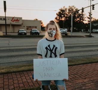 A person is standing on a brick sidewalk, holding a sign that reads 'JUSTICIA PARA VANESSA.' They are wearing a mask and a New York Yankees T-shirt. Behind them, there is a road with parked cars, and a store with a sign that says 'Locke Supply' is visible. The sky appears to be at sunset, with trees and streetlights in the background.