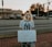 A person is standing on a brick sidewalk, holding a sign that reads 'JUSTICIA PARA VANESSA.' They are wearing a mask and a New York Yankees T-shirt. Behind them, there is a road with parked cars, and a store with a sign that says 'Locke Supply' is visible. The sky appears to be at sunset, with trees and streetlights in the background.
