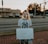 A person is standing on a brick sidewalk, holding a sign that reads 'JUSTICIA PARA VANESSA.' They are wearing a mask and a New York Yankees T-shirt. Behind them, there is a road with parked cars, and a store with a sign that says 'Locke Supply' is visible. The sky appears to be at sunset, with trees and streetlights in the background.