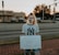 A person is standing on a brick sidewalk, holding a sign that reads 'JUSTICIA PARA VANESSA.' They are wearing a mask and a New York Yankees T-shirt. Behind them, there is a road with parked cars, and a store with a sign that says 'Locke Supply' is visible. The sky appears to be at sunset, with trees and streetlights in the background.