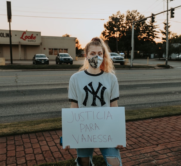 A person is standing on a brick sidewalk, holding a sign that reads 'JUSTICIA PARA VANESSA.' They are wearing a mask and a New York Yankees T-shirt. Behind them, there is a road with parked cars, and a store with a sign that says 'Locke Supply' is visible. The sky appears to be at sunset, with trees and streetlights in the background.