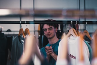 A person is standing behind a clothing rack with various garments on hangers, including a white T-shirt with red text. The individual appears to be in a clothing store, looking towards the camera with hands clasped in front.