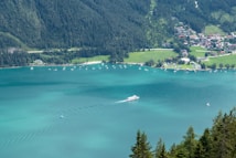 A serene lake with clear turquoise water is surrounded by lush green forests and a small town along the shoreline. A large white boat sails across the center of the lake, leaving gentle ripples in its wake. Several other small boats and sailboats dot the lake, adding a sense of leisure. The backdrop consists of dense forested hills, while the town features neatly arranged houses and a camping area.