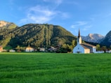 A scenic landscape featuring a white church with a tall steeple in the forefront. The church is situated on a vast, well-manicured grassy field. In the background, there are several houses surrounded by lush green trees. A large, forested mountain rises majestically behind the houses, with power lines or ski lift lines running vertically up the mountain. The sky above is clear with a few wispy clouds.