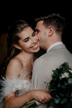 A joyful couple in an intimate embrace, with the man whispering into the woman’s ear as she smiles brightly. They are dressed in formal attire, and the woman holds a bouquet of flowers.