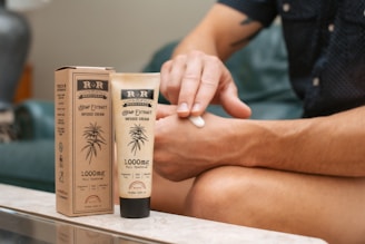 A person is applying hemp extract infused cream to their arm. The product is labeled with 1000mg full spectrum and is packaged in an eco-friendly paper box. The setting appears to be a comfortable indoor environment, with the individual seated on a couch.