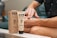 A person is applying hemp extract infused cream to their arm. The product is labeled with 1000mg full spectrum and is packaged in an eco-friendly paper box. The setting appears to be a comfortable indoor environment, with the individual seated on a couch.