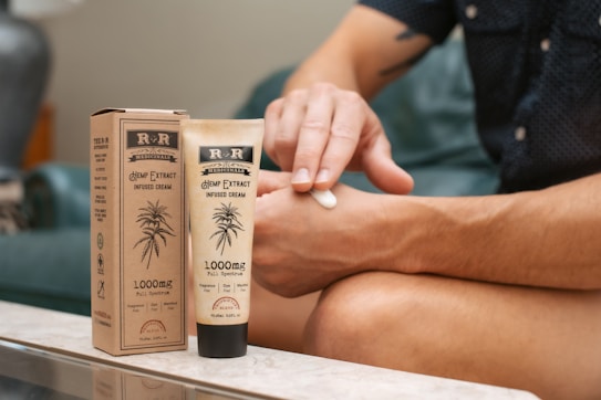 A person is applying hemp extract infused cream to their arm. The product is labeled with 1000mg full spectrum and is packaged in an eco-friendly paper box. The setting appears to be a comfortable indoor environment, with the individual seated on a couch.