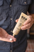 A person holds a tube of hemp extract-infused cream, dispensing a small amount into their other hand. The background is a blurred view of a person wearing a polka-dotted shirt.