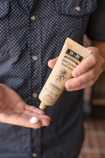 A person holds a tube of hemp extract-infused cream, dispensing a small amount into their other hand. The background is a blurred view of a person wearing a polka-dotted shirt.