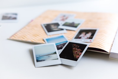 A collection of instant photographs scattered over an open scrapbook with a chevron pattern on the cover. The photos depict various scenes, including landscapes and people, and are casually arranged over a white surface.