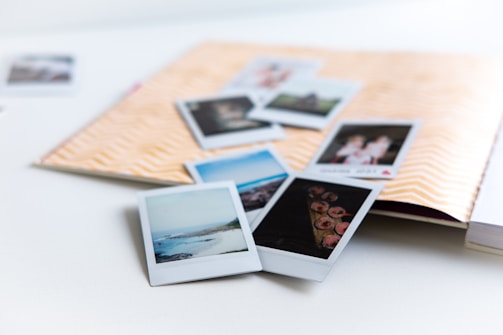 A collection of instant photographs scattered over an open scrapbook with a chevron pattern on the cover. The photos depict various scenes, including landscapes and people, and are casually arranged over a white surface.