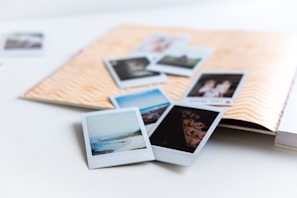 A collection of instant photographs scattered over an open scrapbook with a chevron pattern on the cover. The photos depict various scenes, including landscapes and people, and are casually arranged over a white surface.