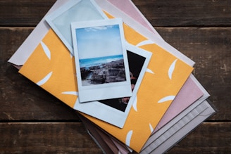 A collection of polaroid photographs is spread across a wooden surface. One of the photos prominently features a scenic ocean view with vibrant blue water and rocky shorelines. These photos rest on top of an assortment of colored and patterned paper, including a notably bright yellow one with white designs.