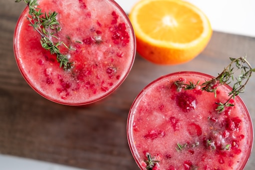 Two glasses filled with a red, fruity beverage topped with fresh thyme sprigs. The drink appears to contain crushed raspberries or a similar berry. In the background, there is a halved orange placed on a wooden surface, adding a fresh and vibrant element to the composition.