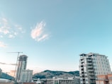 A cityscape featuring modern high-rise buildings under a clear sky with scattered clouds. A construction crane is visible, indicating ongoing development. Hills with sparse vegetation are seen in the background.