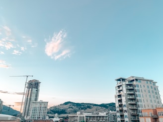 A cityscape featuring modern high-rise buildings under a clear sky with scattered clouds. A construction crane is visible, indicating ongoing development. Hills with sparse vegetation are seen in the background.