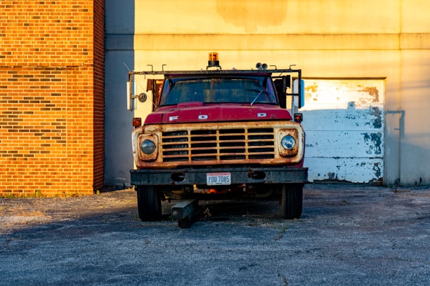 Photograph of a red truck equipped with a custom metal body, parked in an industrial setting with machinery in the background.