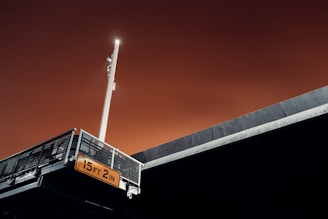 A structure with a metallic grid and a light attached to a pole, illuminated against an ominous orange sky. Below the structure, a clearance sign reads '15 FT 2 IN'. The angles and lighting create a stark and dramatic contrast.