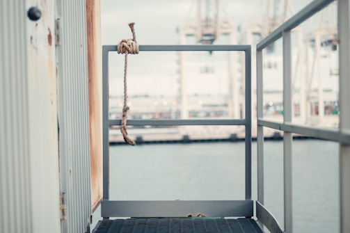 A rope tied to a metal rail on a weathered structure with a view of a port in the background. The scene appears industrial with cranes visible across the water, creating a sense of a dock or harbor.
