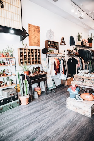 A vintage-themed clothing and decor shop interior with wooden floors and white walls. Clothing is neatly arranged on racks, featuring a mix of shirts, jackets, and hats. The store is decorated with potted plants, including cacti, and various retro items such as posters and signage. There are shelves displaying small potted plants and wooden crates filled with merchandise. A mannequin wearing a vintage t-shirt and sneakers sits on a wooden box.