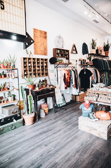 A vintage-themed clothing and decor shop interior with wooden floors and white walls. Clothing is neatly arranged on racks, featuring a mix of shirts, jackets, and hats. The store is decorated with potted plants, including cacti, and various retro items such as posters and signage. There are shelves displaying small potted plants and wooden crates filled with merchandise. A mannequin wearing a vintage t-shirt and sneakers sits on a wooden box.