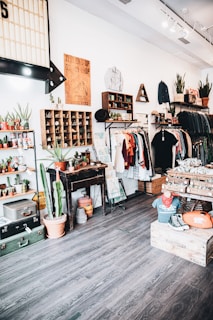 A vintage-themed clothing and decor shop interior with wooden floors and white walls. Clothing is neatly arranged on racks, featuring a mix of shirts, jackets, and hats. The store is decorated with potted plants, including cacti, and various retro items such as posters and signage. There are shelves displaying small potted plants and wooden crates filled with merchandise. A mannequin wearing a vintage t-shirt and sneakers sits on a wooden box.
