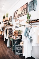 A vintage-style store interior featuring clothing racks with denim and light-colored garments. Shelves display potted plants, leather bags, and various decorative items. A large vintage Levi's sign and pennants are mounted on the wall, along with other unique decor like an old typewriter and a decorative chart with moon phases.