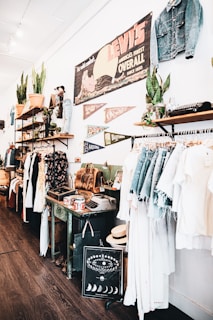 A vintage-style store interior featuring clothing racks with denim and light-colored garments. Shelves display potted plants, leather bags, and various decorative items. A large vintage Levi's sign and pennants are mounted on the wall, along with other unique decor like an old typewriter and a decorative chart with moon phases.